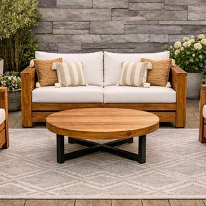 Puede incluir: Juego de muebles de patio exterior de madera con cojines blancos y cojines decorativos. Incluye un sofá, dos sillones y una mesa de centro redonda con base negra. El conjunto está sobre una alfombra estampada con una pared de piedra de fondo.
