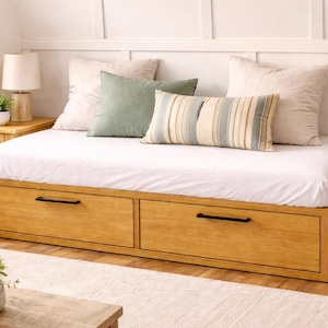 May include: A wooden daybed with two drawers, a white mattress, and decorative pillows. The bed frame is light brown with black handles. The pillows are cream, green, and striped. A small side table with a lamp and a plant is visible.
