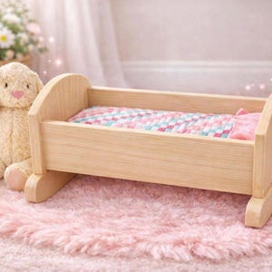 Könnte beinhalten: Ein hölzernes Puppenbett mit einer rosa und gemusterten Decke und einem Kissen liegt auf einem rosa Teppich. Ein Stoffhase mit langen Ohren und einer rosa Nase befindet sich neben dem Bett. Das Bett ist aus hellem Holz.