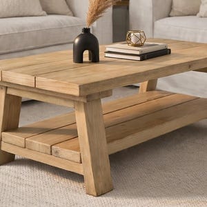 May include: A light brown wooden coffee table with a lower shelf. The table has a rectangular top and sturdy legs. A black vase, books, and a gold candle holder are on the table. The table is in a living room setting.