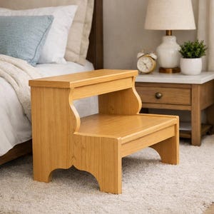 May include: A two-step wooden step stool with a light oak finish. The stool has a rectangular top step and a lower step, with curved side supports. It is placed on a light-colored carpet, near a bed and nightstand.