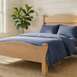 May include: A wooden bed frame with a curved headboard and footboard. The bed is made up with a navy blue duvet cover and matching pillows. The room has a neutral color palette with a large window and potted plants.