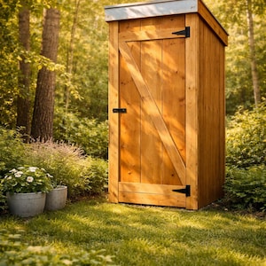 May include: A wooden outhouse with a slanted roof and a door with a diagonal brace. The outhouse is made of light-colored wood and has black hinges and a latch. It stands in a grassy area surrounded by trees and plants.