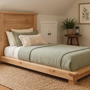 May include: A light-colored wooden bed frame with a headboard, featuring a sage green quilt and pillows. The bed sits on a woven rug, creating a cozy bedroom setting. A framed botanical print hangs on the wall.