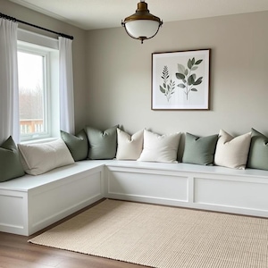 Puede incluir: Un banco blanco empotrado con cojines verdes y crema. Una impresión botánica enmarcada cuelga de la pared sobre el banco. Una alfombra beige está en el suelo frente al banco, y cortinas blancas enmarcan una ventana.