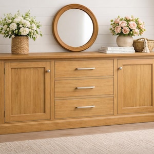 May include: A light oak wood sideboard with three drawers and two cabinet doors. A round mirror hangs above the sideboard. Decorative elements include floral arrangements in vases and baskets.