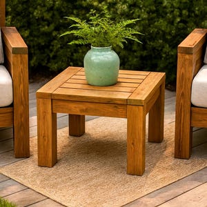 Puede incluir: Juego de muebles de patio al aire libre con dos sillones de madera con cojines color crema y una mesa auxiliar cuadrada de madera. Un jarrón verde con vegetación se encuentra sobre la mesa. El conjunto está sobre una alfombra tejida.