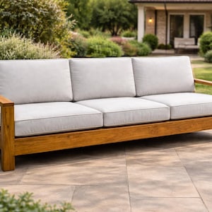 Puede incluir: Un sofá de exterior de tres plazas con cojines de color gris claro y una estructura de madera. El sofá es de madera y tiene forma rectangular. Los cojines son de color gris claro con un ribete gris más oscuro. El sofá está sobre un patio de piedra.