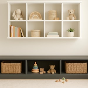 May include: A white wall-mounted shelf unit with eight cubbies, above a dark gray storage bench. The shelves display toys, books, and decorative items, including a stuffed bunny, a wooden rainbow, and a teddy bear. The bench has three storage compartments.