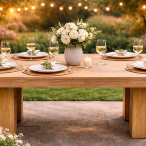 Könnte beinhalten: Ein Holztisch für ein Essen im Freien. Der Tisch ist mit weißen Tellern, Weingläsern und Blumengestecken gedeckt. Eine Kerze und Lichterketten tragen zum Ambiente bei.