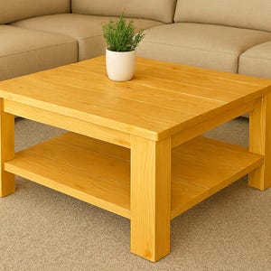 Op de afbeelding: Een vierkante, lichtgekleurde houten salontafel met een onderste plank. De tafel heeft een natuurlijke houtnerf afwerking en is ongeveer 91 cm vierkant. Een kleine plant in een pot staat op de tafel.