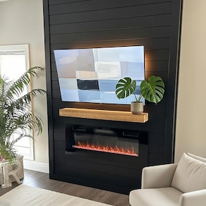 May include: A modern black fireplace and entertainment centre with a mounted TV and a wooden shelf. A potted plant sits on the shelf. The fireplace has a simulated flame effect. The wall is painted black.