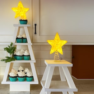 May include: A white wooden Christmas tree-shaped display stand with teal cupcake holders and white frosting. A lighted yellow star sits atop the tree. Another stand holds a yellow star on a spring.