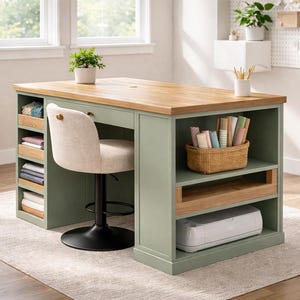 May include: A sage green and wood-toned craft table with a beige upholstered chair. The table has open shelving and a built-in drawer. A white printer sits on the bottom shelf. A small potted plant and a basket of craft supplies are visible.