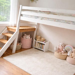 May include: A white loft bed with a wooden staircase and a small desk underneath. The room is decorated with stuffed animals, a pink basket, and a small bookshelf. A cream-colored rug covers the floor.