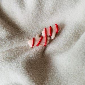 May include: A small, abstract, handmade polymer clay pin. The pin is a light pink color with red stripes. The pin is on a white, textured surface.