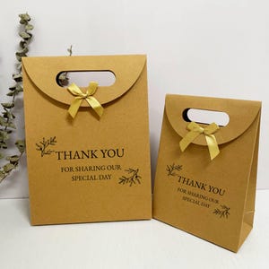 Könnte beinhalten: Zwei braune Geschenkpapierbeutel mit einem Griffausschnitt und einer goldenen Schleife. Die Beutel tragen den schwarzen Aufdruck "THANK YOU FOR SHARING OUR SPECIAL DAY" sowie Blattillustrationen. Eine Tasche ist größer als die andere.
