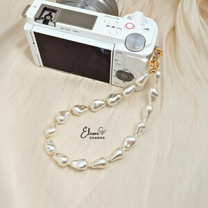 May include: A white digital camera with a pearl wrist strap. The strap features irregular, off-white pearls with gold-colored accents. The camera is on a cream-colored fabric. The brand name "Elesar CHARMS" is visible.