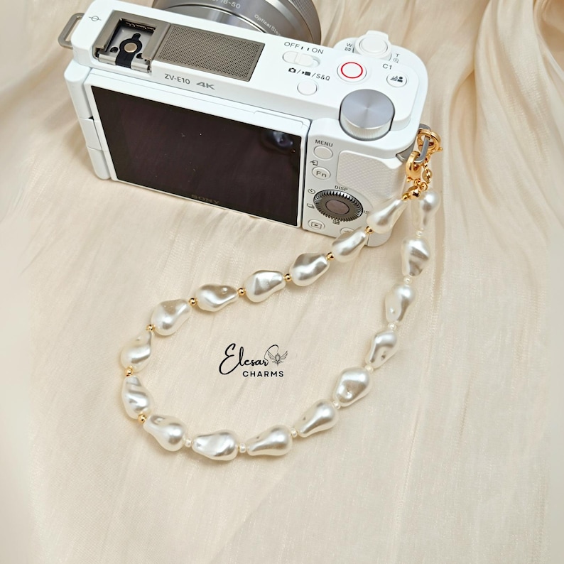 May include: A white digital camera with a pearl wrist strap. The strap features irregular, off-white pearls with gold-colored accents. The camera is on a cream-colored fabric. The brand name "Elesar CHARMS" is visible.