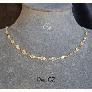 May include: A gold-toned chain necklace featuring a series of clear, oval-shaped cubic zirconia stones linked together. The necklace displays the text "Elesar Atelier" and "Oval CZ" in white lettering against the grey display bust.