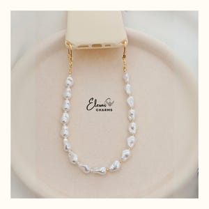 May include: A cream-colored phone case with a gold-colored chain of irregular white pearls. The chain is attached to the phone case with gold-colored clasps. The brand name "Elesar Charms" is printed on the image.