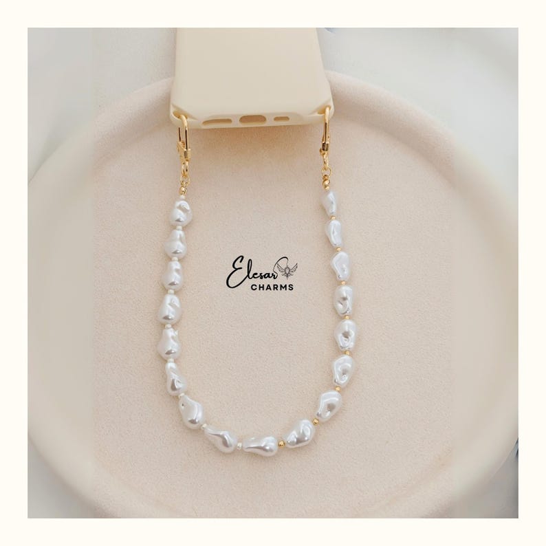 May include: A cream-colored phone case with a gold-colored chain of irregular white pearls. The chain is attached to the phone case with gold-colored clasps. The brand name "Elesar Charms" is printed on the image.