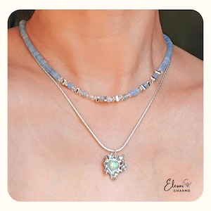 May include: A two-strand necklace set. The top strand is a light blue beaded choker. The second strand is a silver chain with a heart-shaped pendant with a white and green stone. The pendant is surrounded by a silver floral design.