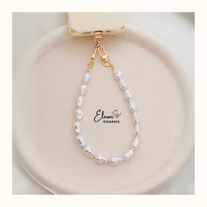 May include: A white pearl phone charm with gold-colored accents. The charm is attached to a cream-colored phone case. The charm is made of irregular-shaped pearls and small gold beads. The brand name "Elesar CHARMS" is printed on the image.