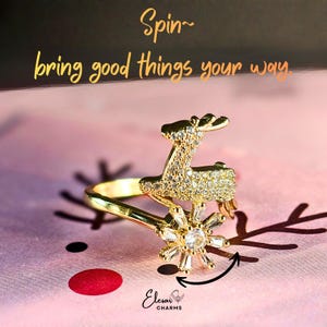 Peut inclure: Bague dorée ornée d'un renne et d'un flocon de neige rotatif serti de cristaux. La bague est présentée sur un fond rose avec le texte "Spin~ bring good things your way". Un bijou festif.
