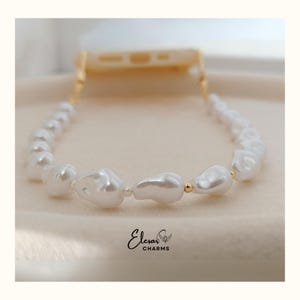 May include: A white pearl phone strap with gold-colored accents. The strap is made of irregular-shaped pearls and connects to a phone case. The brand name "Elesar Charms" is visible in the bottom center.