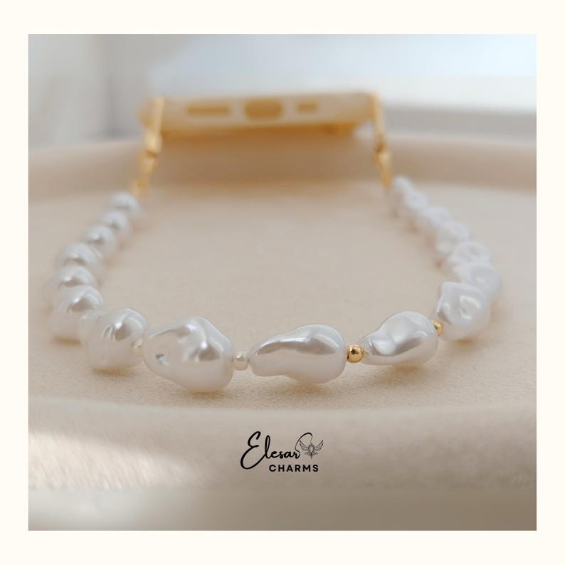 May include: A white pearl phone strap with gold-colored accents. The strap is made of irregular-shaped pearls and connects to a phone case. The brand name "Elesar Charms" is visible in the bottom center.