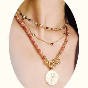 Peut inclure: Ensemble de colliers superposés. La chaîne supérieure est ornée de petites perles colorées. La chaîne du milieu est une fine chaîne dorée avec des breloques en forme de cœur. La chaîne inférieure est composée de grosses perles roses et d'un pendentif doré.