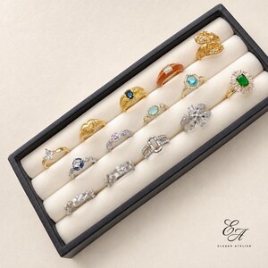 Op de afbeelding: Een collectie van dertien diverse ringen gepresenteerd in een zwarte rechthoekige sieradenlade met witte fluwelen gleuven. De ringen hebben verschillende ontwerpen, waaronder goud- en zilverkleurige banden, gekleurde edelstenen en heldere kristallen.