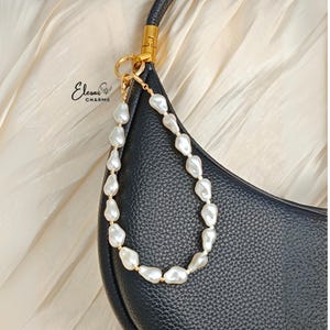 May include: A black leather handbag with a gold-tone handle and a pearl beaded strap. The strap is made of irregular white pearls strung together with gold-colored beads. The brand name "Elesar Charms" is visible in the upper left corner.