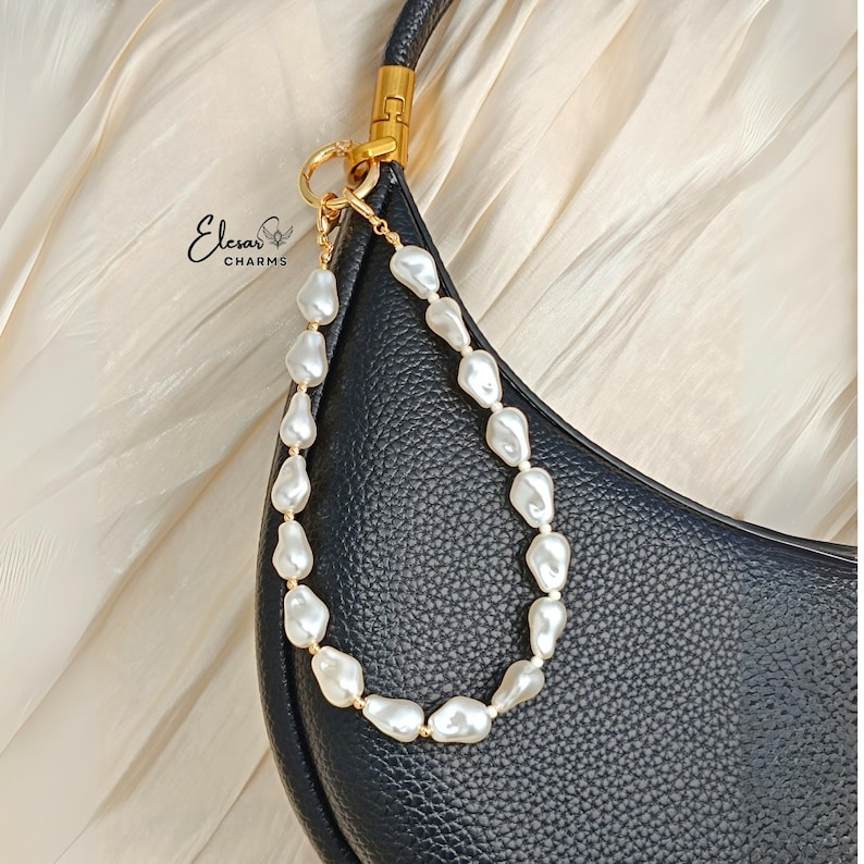 May include: A black leather handbag with a gold-tone handle and a pearl beaded strap. The strap is made of irregular white pearls strung together with gold-colored beads. The brand name "Elesar Charms" is visible in the upper left corner.