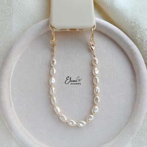 Pode incluir: Um conector de pulseira de relógio cor de creme preso a uma corrente dourada com pérolas brancas ovais. A corrente é fixada com fechos lagosta dourados. O texto Elesar CHARMS está impresso em preto perto do centro da corrente.