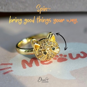 May include: A gold-colored ring featuring a cat face design encrusted with small, sparkling stones. The ring has a smooth band and the cat's face has black eyes and detailed features. Text on the image reads "Spin~ bring good things your way."