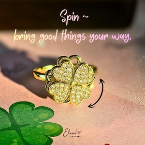 Op de afbeelding: Een goudkleurige ring met een klaverbladontwerp met vier bladeren, elk blad bezet met kleine, sprankelende stenen. De tekst "Spin ~ bring good things your way" staat op een roze achtergrond.