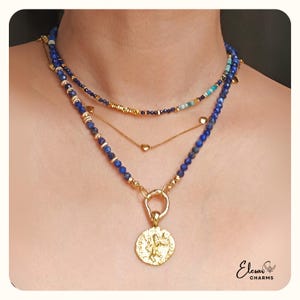 Peut inclure: Un ensemble de colliers superposés avec des perles de lapis-lazuli, des chaînes dorées et un pendentif pièce de monnaie en or. Le collier comprend différentes tailles de perles et longueurs de chaînes, créant un look multidimensionnel.