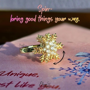 May include: A gold-colored ring with a snowflake design, encrusted with small, clear gemstones. The ring is on a pink surface with the text "Spin~ bring good things your way." and "Unique, just like you."