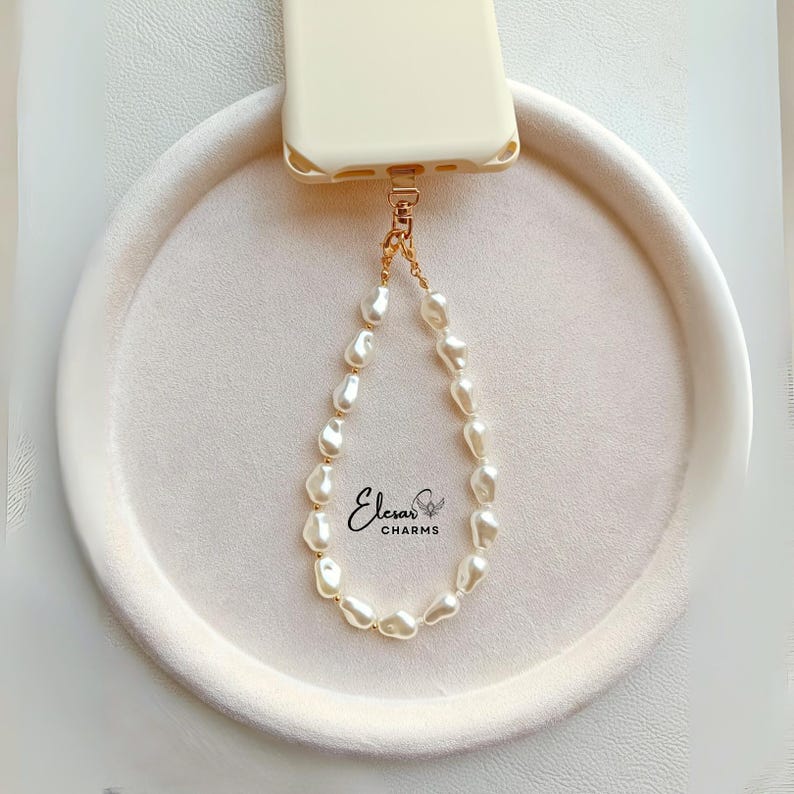 May include: A cream-colored phone case with a gold-toned attachment holding a pearl phone charm. The charm is made of irregular, off-white pearls strung together with a gold chain. The brand name "Eleras Charms" is printed in cursive.