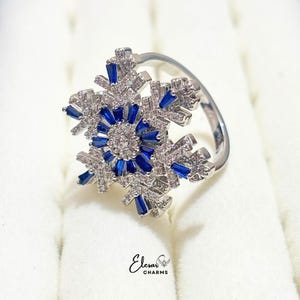 Peut inclure: Une bague argentée avec un grand motif central en forme de flocon de neige. Le design est incrusté de nombreuses petites pierres claires et accentué par des pierres bleu foncé taille baguette disposées en rayon. Le texte Elesar CHARMS est visible.