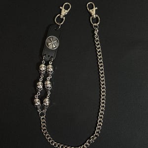 May include: A silver-toned wallet chain with a black leather strap and two lobster claw clasps. The chain features a decorative metal cross, a series of skull charms, and a curb chain.