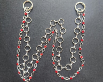 May include: Silver-toned chain accessory with red gemstone accents. The chain features large circular links and smaller chains with red stones. Two large circular clasps are at the top.