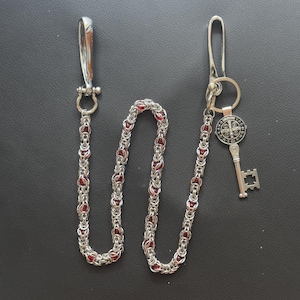 Może przedstawiać: Srebrny łańcuch do portfela z czerwonymi akcentami. Łańcuch jest połączony ze srebrnym zapięciem i brelokiem z kluczem i medalionem. Medalion ma wzór krzyża. Łańcuch ma długość około 60 cm.