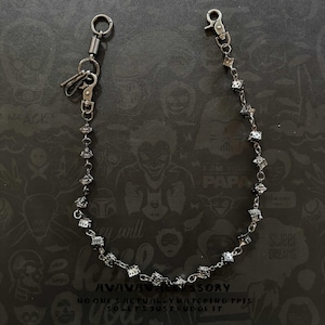 Op de afbeelding: Een zilverkleurige portemonnee ketting met een veerklem, sleutelring en karabijnhaak. De ketting is versierd met vierkante, facetgeslepen versieringen. De achtergrond heeft een donker patroon met de tekst "AVAVAV ACCESSORY".