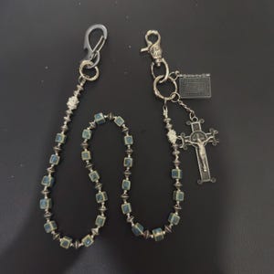 Può includere: Una catena per portafoglio argentata con un design di perline blu e argento. Un'estremità presenta un fermaglio, mentre l'altra ha un ciondolo a forma di croce e un piccolo ciondolo rettangolare. La catena è progettata per essere attaccata a un portafoglio o alle chiavi.