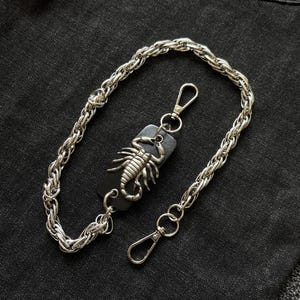 Pode incluir: Corrente de metal prateado com fecho mosquetão e um pendente decorativo de escorpião em suporte de couro preto. A corrente é de estilo trançado espesso, e o escorpião é detalhado.