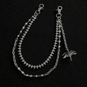 May include: Silver chain accessory with two lobster claw clasps. The chains have cross and pearl accents, with a dangling winged figure charm. The accessory is set against a black background.