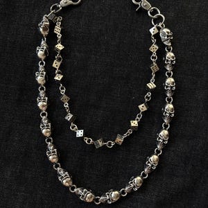 Pode incluir: Corrente de carteira prateada com pingentes de caveira e dados. A corrente tem duas pontas, cada uma com pingentes de caveira e dados pequenos. Um acessório gótico.